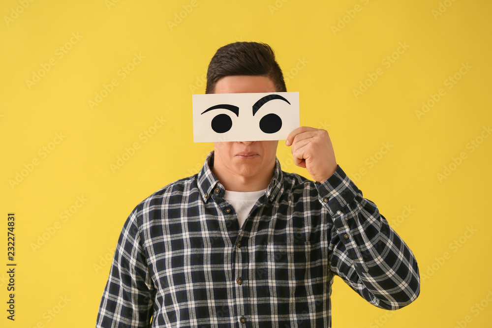 Emotional young man hiding face behind sheet of paper with drawn eyes on color background Emotional young man hiding face behind sheet of paper with drawn eyes on color background