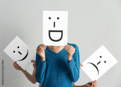Young women hiding faces behind sheets of paper with drawn emoticons on light background photo