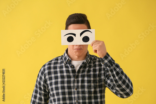 Emotional young man hiding face behind sheet of paper with drawn eyes on color background