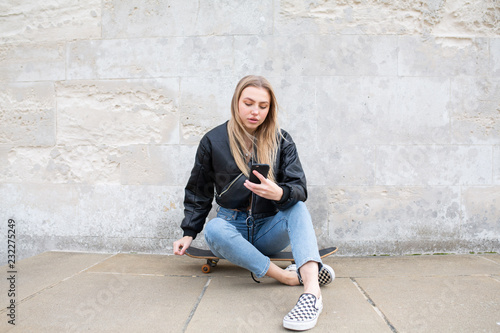 Teenage girl sitting on her skate board texting outside photo