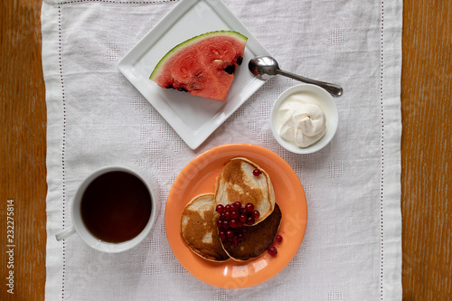 Top view of pancakes in orange plate and glass of sourcream with red berries, slice of watermelon, wooden table and white cloth. photo