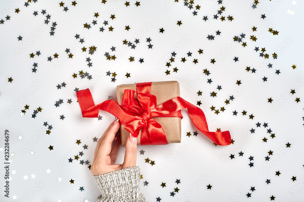 Female hands holding present with red bow on white background with delicate wavy ribbons and metallic star shaped confetti. Flat lay style Female hands holding present with red bow on white background with delicate wavy ribbons and metallic star shaped confetti. Flat lay style