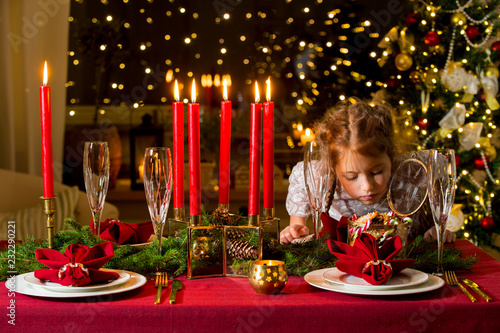 Cute little girl celebrating Christmas, taking candies and gingerbread from glass jar and eating. Served table with candles, Red tablecloth and napkins, white china, gold cutlery, crystal glasses.