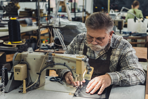 serious mature male tailor in apron and eyeglasses working on sewing machine at studio