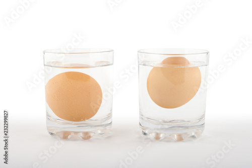 Two glasses of water with a fresh egg on the left and a rotten egg on the right side on white background. Bad egg floats in water. photo