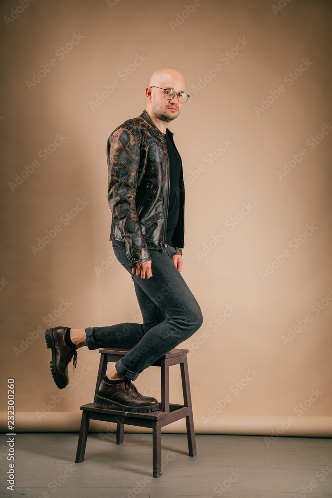 Lifestyle portrait of fashionable stylish bald man in glasses. Awesome man  wearing biker leather jacket, black jeans, oxford shoes. Cool handsome well  dressed boy on coffee paper studio background. Stock Photo