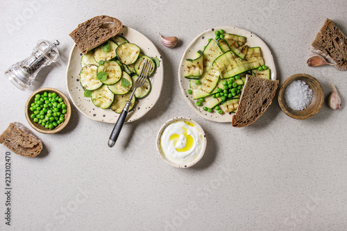 Variety of grilled zucchini salad with green pea, yogurt dip, garlic and rye sliced bread in spotted ceramic plates over grey background. Vegetarian food. Flat lay, space