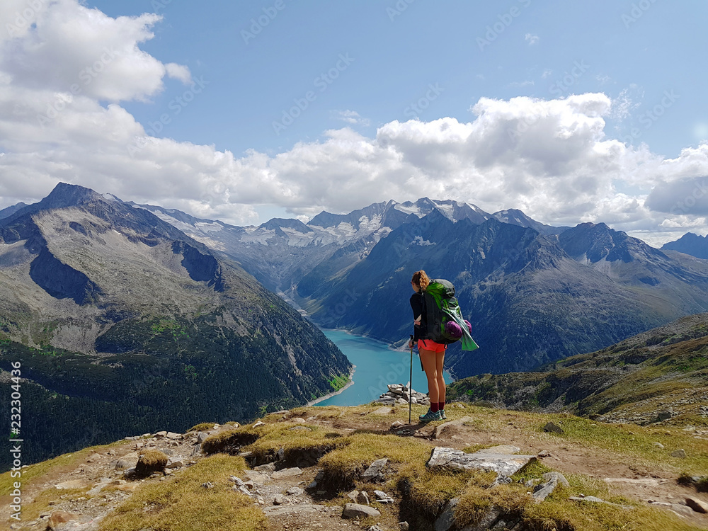 Hiker with backpack relaxing on top of a mountain and enjoying valley view with lake during trip in the alps