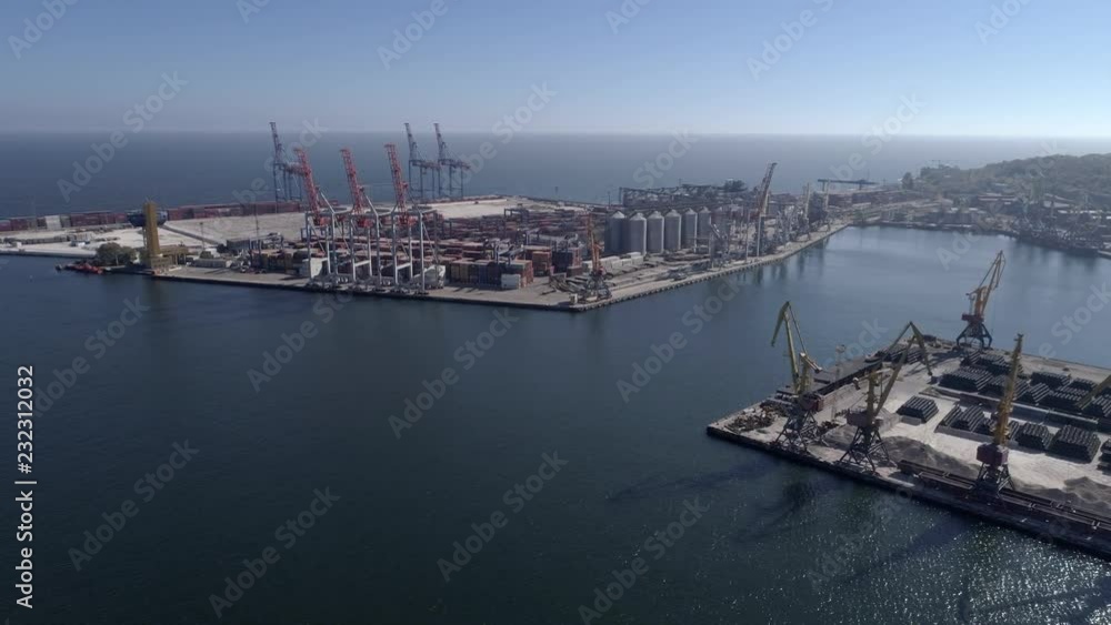 Flying above Industrial trading port with grain storage elevator with containers and lifting cranes for loading and unloading of vessel on Sea Embankment against blue sky and shiny water, aerial view Flying above Industrial trading port with grain storage elevator with containers and lifting cranes for loading and unloading of vessel on Sea Embankment against blue sky and shiny water, aerial view