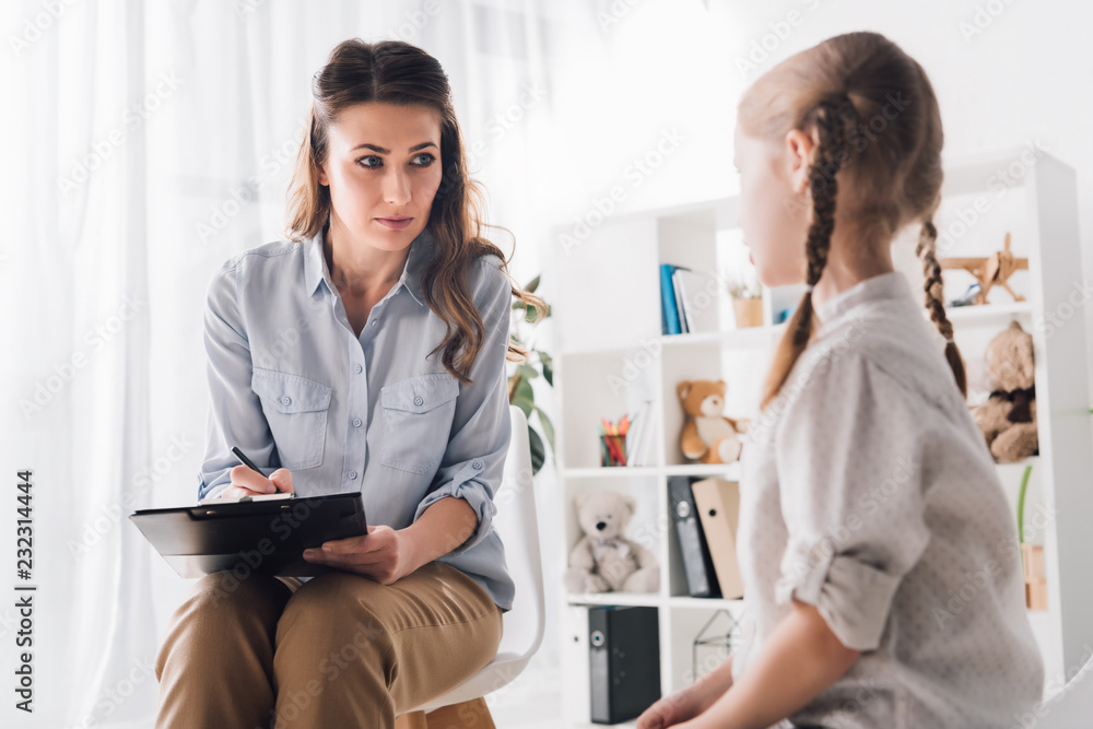 Fototapeta premium adult psychologist with clipboard talking to little child in office