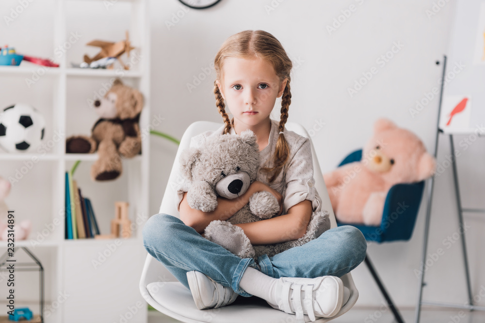 lonely little child sitting on chair and embracing her teddy bear while looking at camera lonely little child sitting on chair and embracing her teddy bear while looking at camera