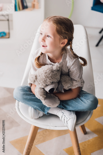 happy little child sitting on chair and embracing her teddy bear