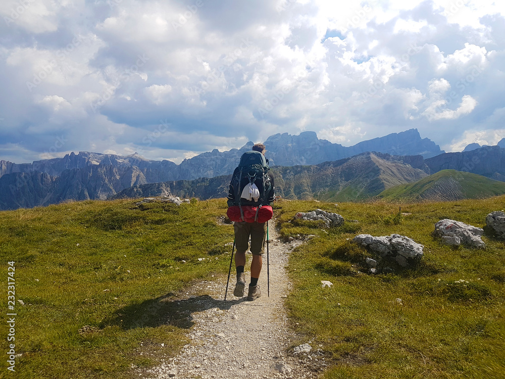 Hiker with backpack walking a trail on top of a mountain and enjoying valley view during trip in the alps