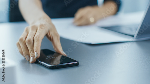 Shot of the Businesswoman Working at Her Office Desk and Reaching Out for Her Smartphone. Woman Picks up Mobile Phone from Her Desk. Focus on a Phone.