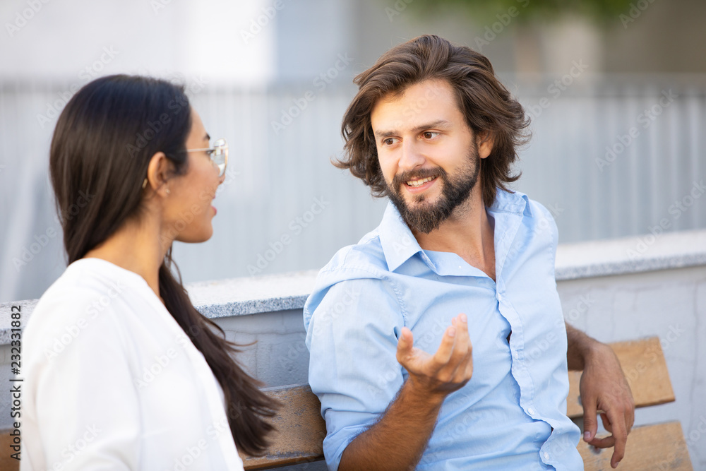 Obraz premium Cheerful handsome man gesturing and talking to colleague while sitting on bench in city. Professional stylish couple on meeting outdoors. Teamwork concept