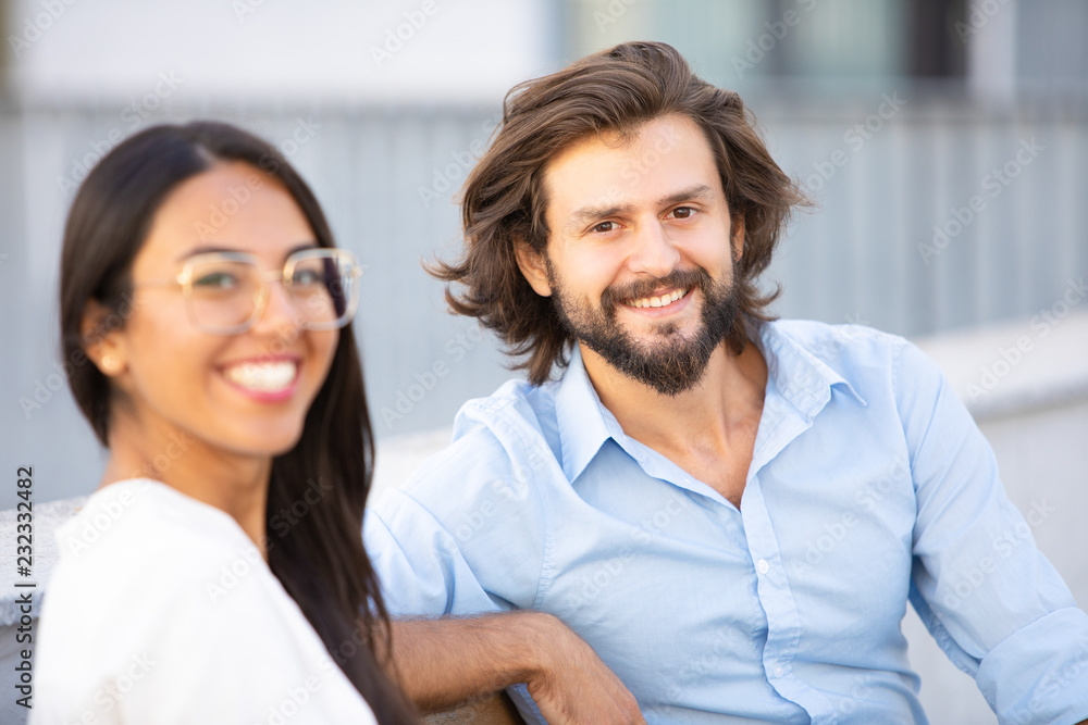 Smiling multiethnic friends looking at camera outdoors. Happy latin woman and handsome cheerful man sitting on bench in city. Friendship concept