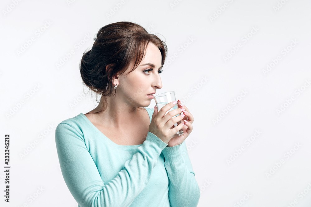 Beautiful girl with brown-haired woman dressed mint-colored sweater on a light background holds a glass of pure drinking water looks to the left Beautiful girl with brown-haired woman dressed mint-colored sweater on a light background holds a glass of pure drinking water looks to the left