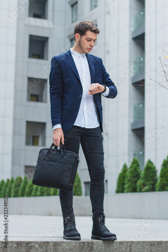 Business style. Young businessman in his hand the briefcase. The guy in the blue jacket looks at his watch.