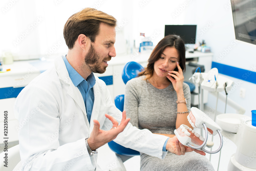 Obraz premium Attractive young happy smiling dentist in dentist office explaining his female patient the teeth model denture and showing the procedure of dental teeth implants.