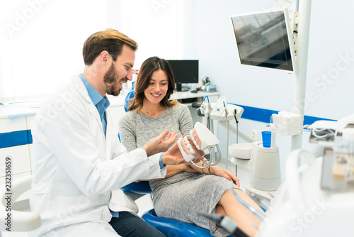 Happy smiling dentist and beautiful woman patient looking at teeth model dentures explaining procedure of dental teeth implants in the modern dental clinic. Dentistry concept. 