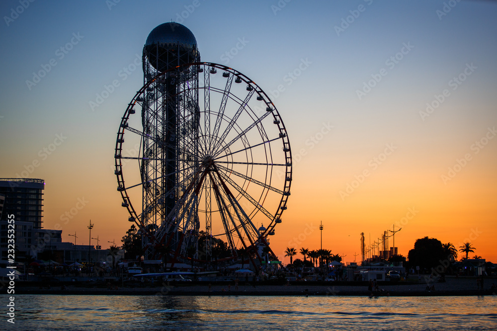 Ferris wheel and Georgian alphabet tower on orange sunset background. Silhouettes of the wheel and tower in the evening in the lights of illumination with the reflection of light in the water.