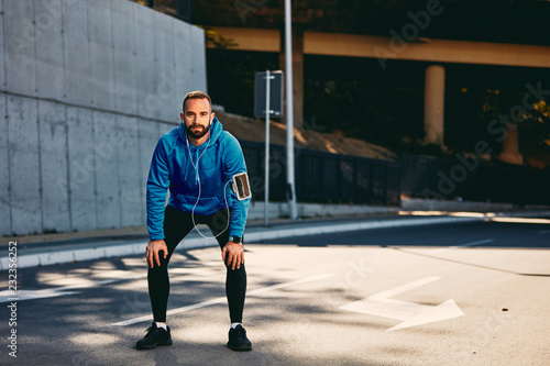 Young Caucasian bearded man standing on a street and resting from running while looking at camera. Sportswear on, earphones in ears. Healthy lifestyle concept. © chika_milan