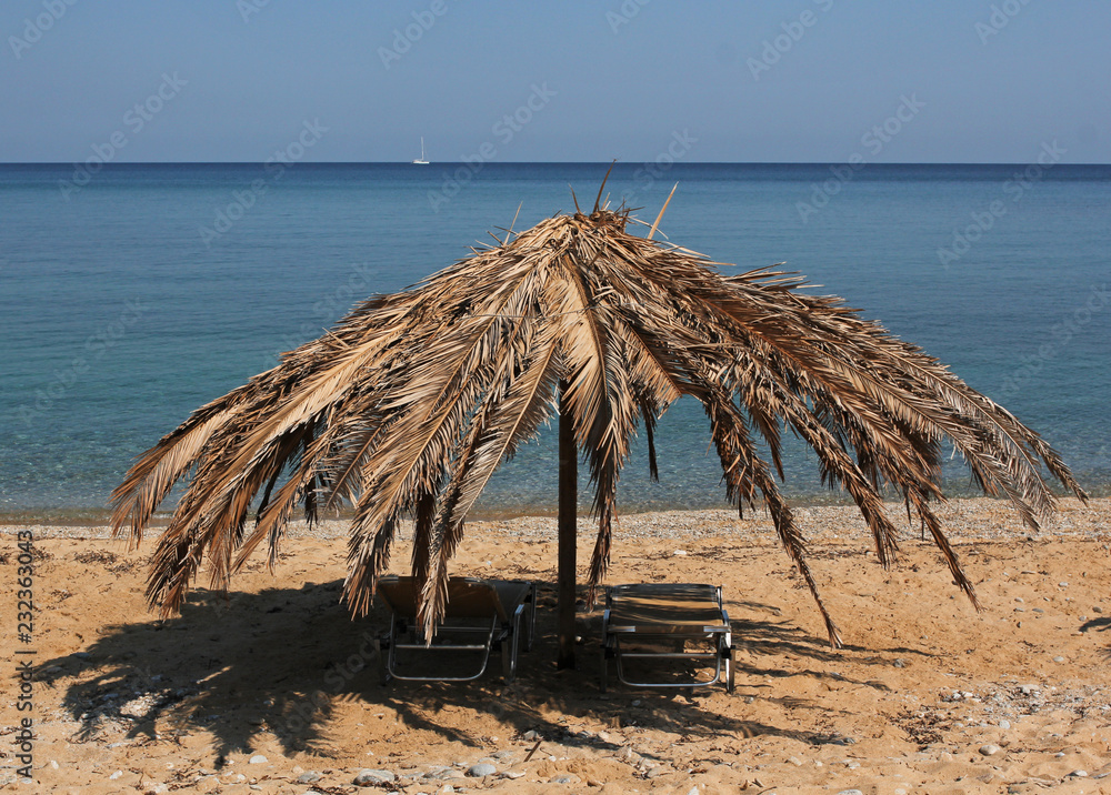 Palm leaves umbrella on the sandy beach, sailboat on blue sea in background Palm leaves umbrella on the sandy beach, sailboat on blue sea in background