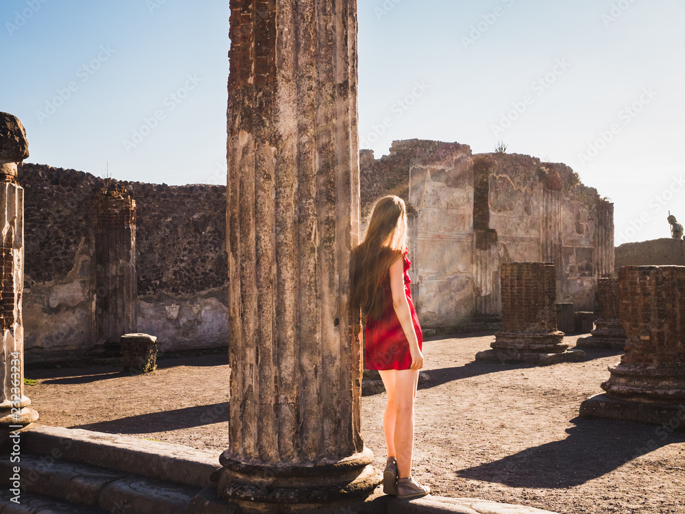 Φωτογραφία Stylish woman in a red dress looking into the distance against the backdrop of ancient ruins and the sun's rays of the evening sun