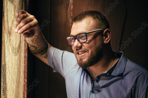 brutal bearded man with a tattoo on his arm, a portrait of a man in dramatic light against a brown wooden wall, attractive bearded male with tattoo on arm dressed in a shirt, man looks in the side