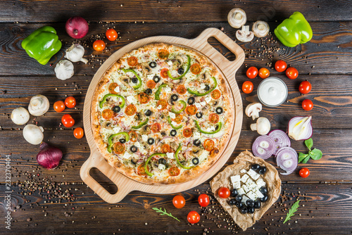 Big pizza with cheese, tomatoes, black olives and paprika on a round cutting board on a dark wooden background. Ingredients.