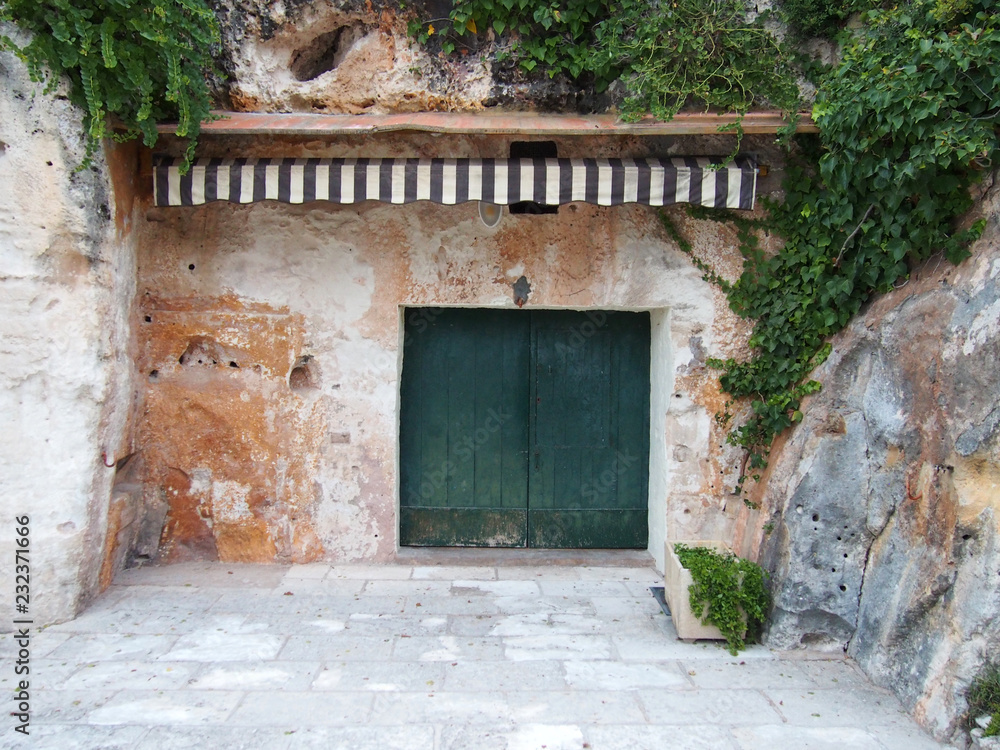 A stone shelter carved out of a cliff with green vegetation wooden doors and a striped awning