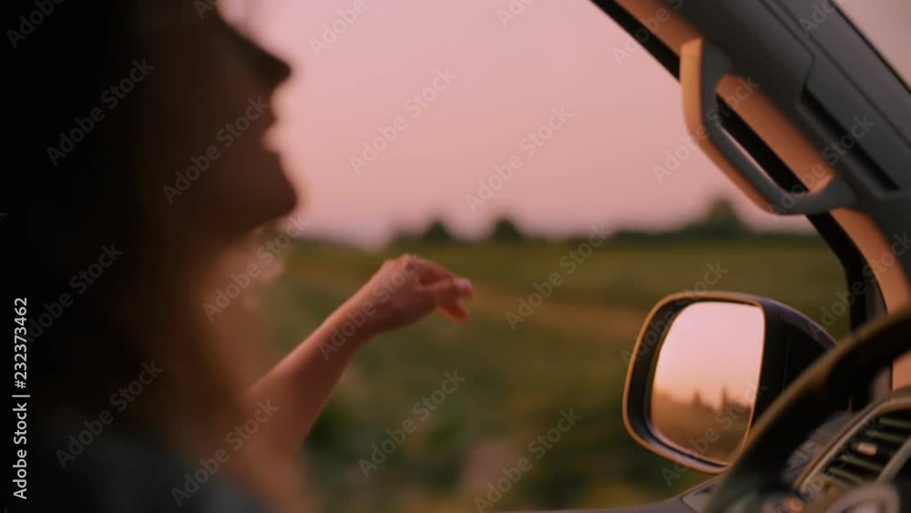 Cinematic inspirational video of young woman travelling by car or camper van, opens window to breathe fresh air of countryside, moves hand in wind. Sings melody of song, summertime vacation vibes