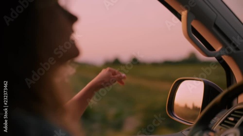 Cinematic inspirational video of young woman travelling by car or camper van, opens window to breathe fresh air of countryside, moves hand in wind. Sings melody of song, summertime vacation vibes