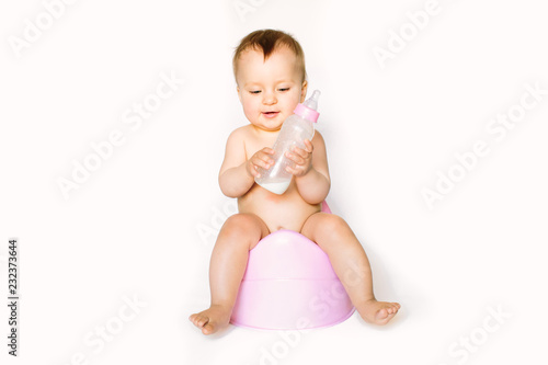 Charming photo of cute baby girl holding a boottle with baby milk and sitting on a pink potty. Isolated on white background
