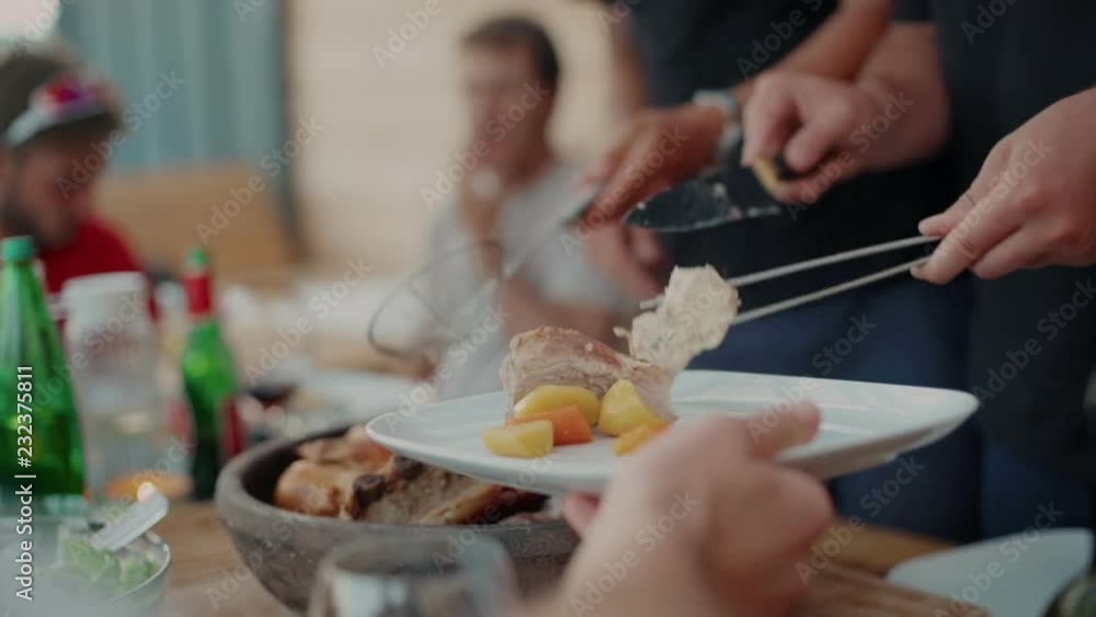 Over shoulder depth of field shot of man reaching with empty plate to be served steaming hot delicious meat dish and boiled vegetables. Friends pass on plates to share and enjoy dinner together Over shoulder depth of field shot of man reaching with empty plate to be served steaming hot delicious meat dish and boiled vegetables. Friends pass on plates to share and enjoy dinner together