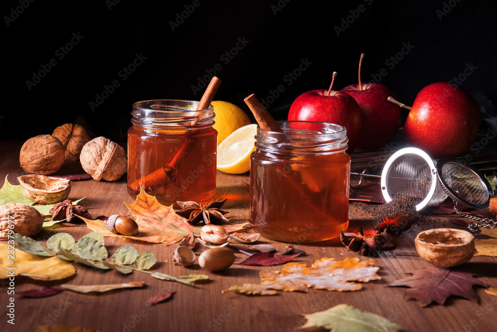 Dark wooden table with 2 glasses of tea with fall, autumn decoration and dark, black background. Colorful leaves, lemon, cinnamon, walnuts, apples, tea, walnut shells, hazelnuts, tea strainer, acorns.