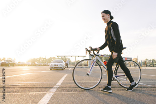 Happy young man in dark sportswear and cap, pulls a bicycle on the street in the background of the sunset and smiles. Cyclist at sunset.