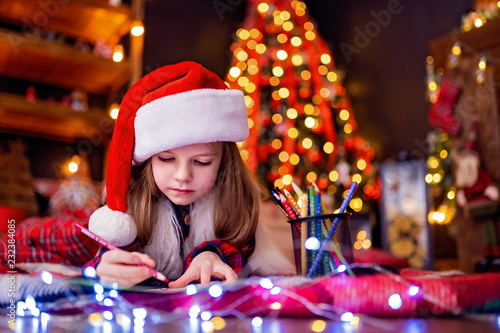 Funny girl in Santa hat writes letter to Santa near christmas tree