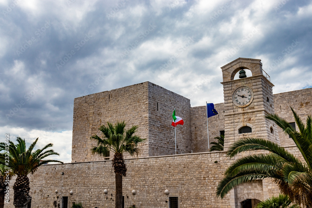 The flags of Italy and of the European community waving on the Swabian castle of Trani. Close-up of the tower with the clock. Stone fortress, on the sea. In Puglia, near Bari, Barletta, Andria.
