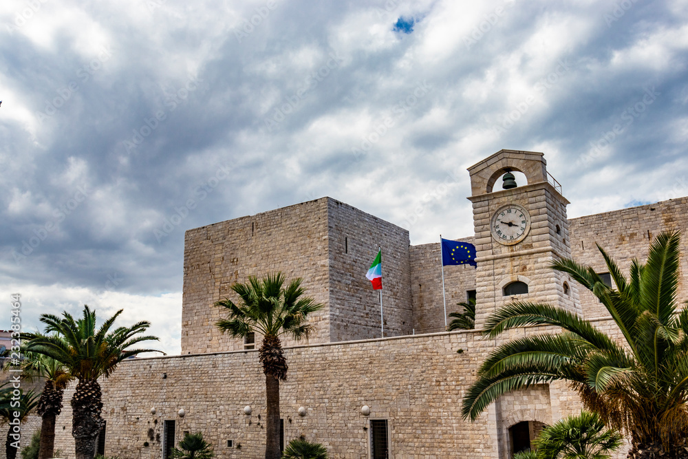 The flags of Italy and of the European community waving on the Swabian castle of Trani. Close-up of the tower with the clock. Stone fortress, on the sea. In Puglia, near Bari, Barletta, Andria. The flags of Italy and of the European community waving on the Swabian castle of Trani. Close-up of the tower with the clock. Stone fortress, on the sea. In Puglia, near Bari, Barletta, Andria.