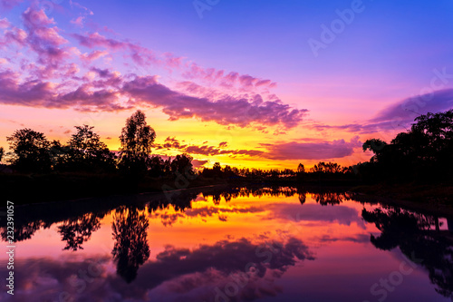 Sun setting with silhouette trees and bush around the big pond in rural of Thailand, shot taken by using long exposure therefore smooth water surface like mirror