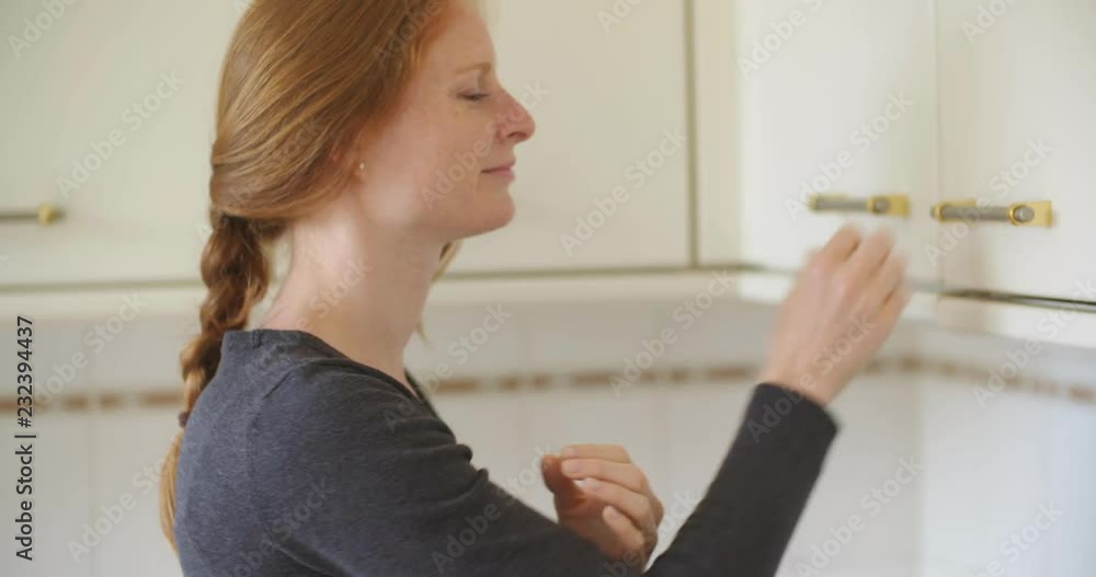 An attractive redhead young woman smiles at the camera after closing a kitchen cupboard. An attractive redhead young woman smiles at the camera after closing a kitchen cupboard.