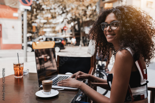Smiling young ravishing Brazilian businesswoman in eyeglasses and with a gorgeous curly bulky hair is sitting in a street bar, working on her laptop and drinking delicious cappuccino from the cup near