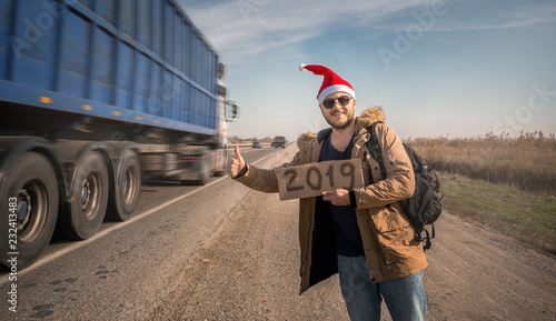 The concept of a new year and journey. A young man hitchhiking, holding a sign that says "2019", dressed in a down jacket, glasses and Santa hat.