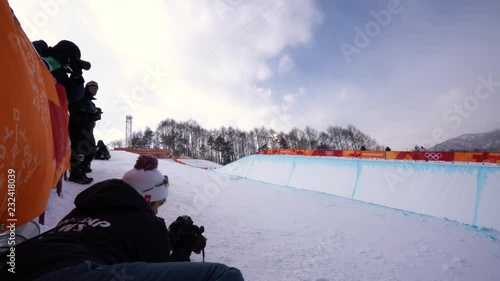 Steady close up shot of a snow boarding competetion on an afternoon at the Pyeongchang Winter Olympics and an athlete performing freestyle tricks, taken from the platform beside the snowboard track