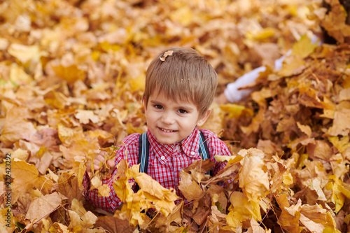 Child boy is lying and playing on fallen leaves in autumn city park.