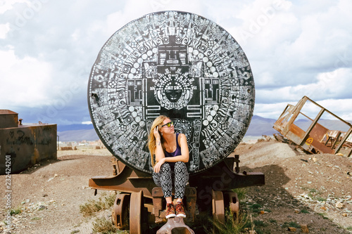 Bolivia. Cemetery of old trains. The blonde girl sits on a train car in sunglasses. Clear day. The train car is painted with an unusual black-and-white pattern in a mazaic style. photo