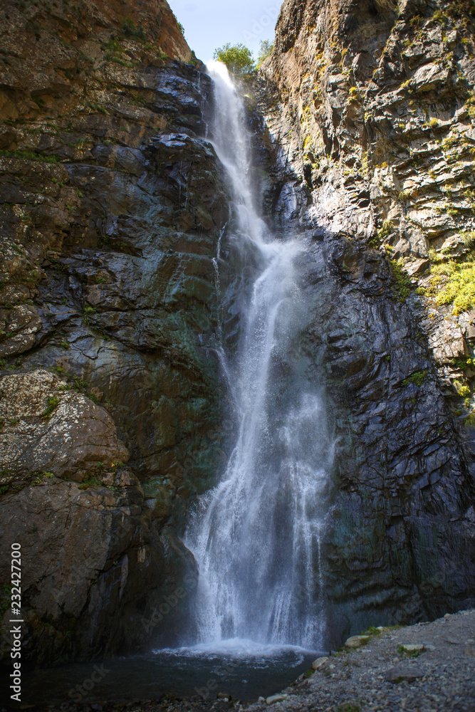 Obraz premium Gveleti waterfall in Georgia. The stream of water pouring from the top on the wet gray rocks, the sunlight playing in the spray of water and is reflected on the wet black rocks.