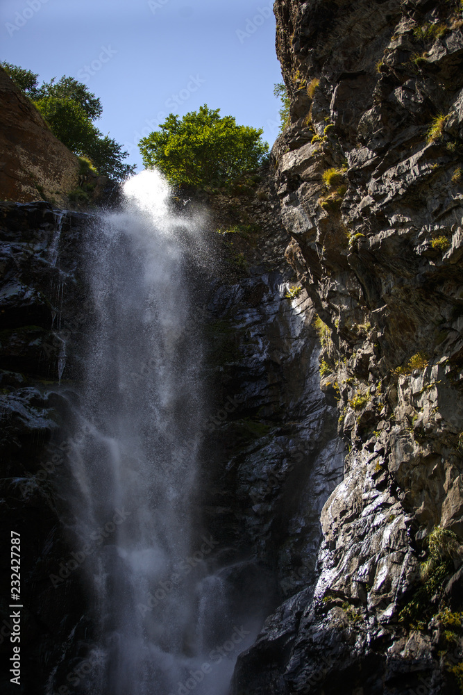 Obraz premium Gveleti waterfall in Georgia. The stream of water pouring from the top on the wet gray rocks, the sunlight playing in the spray of water and is reflected on the wet black rocks.