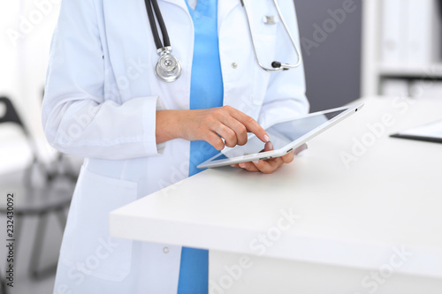 Close-up of female physician using digital tablet while standing near reception desk at clinic or emergency hospital. Unknown doctor woman at work. Medicine and healthcare concept